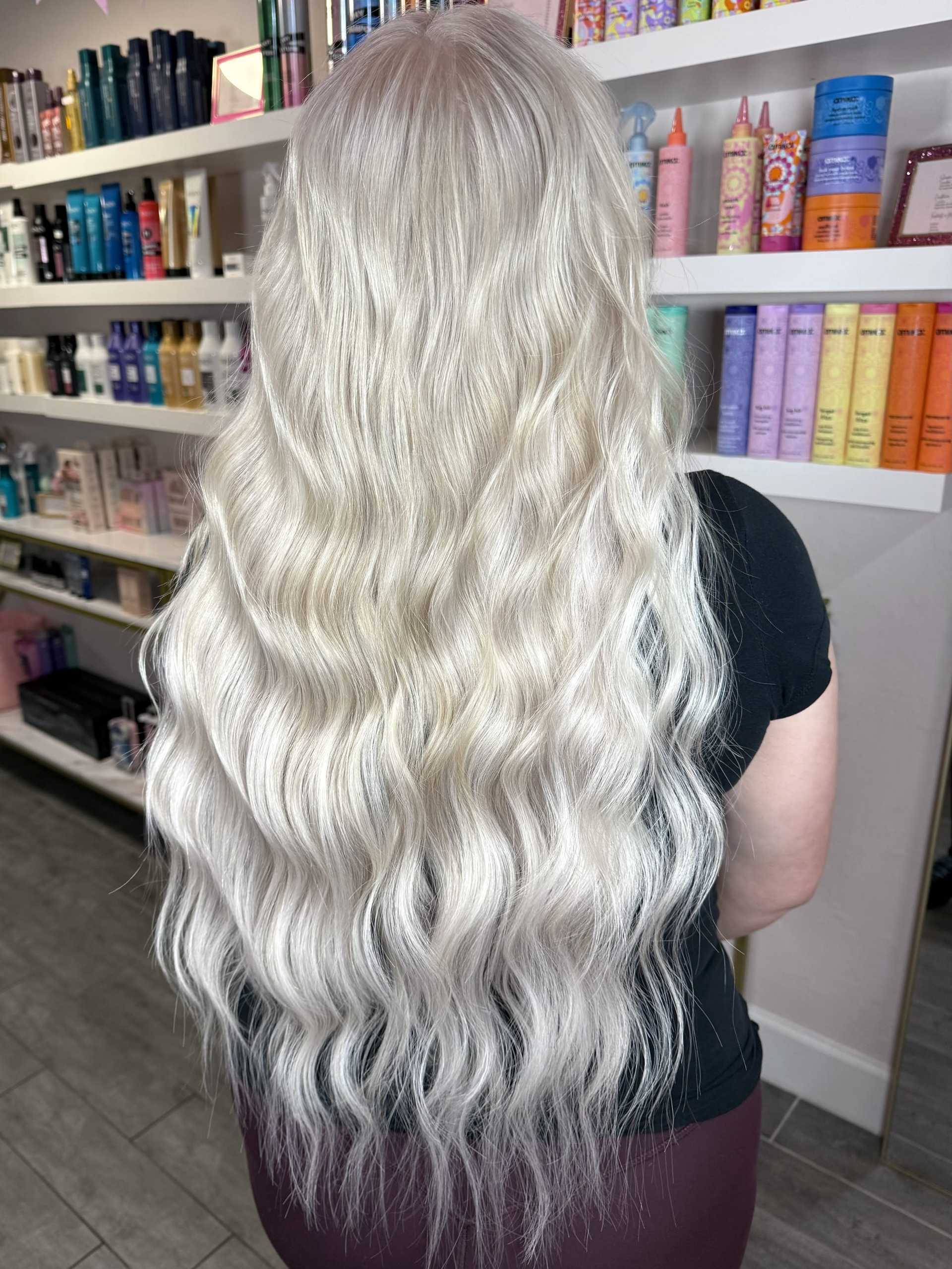 Long wavy platinum blonde hair in a salon with colorful hair products on shelves.